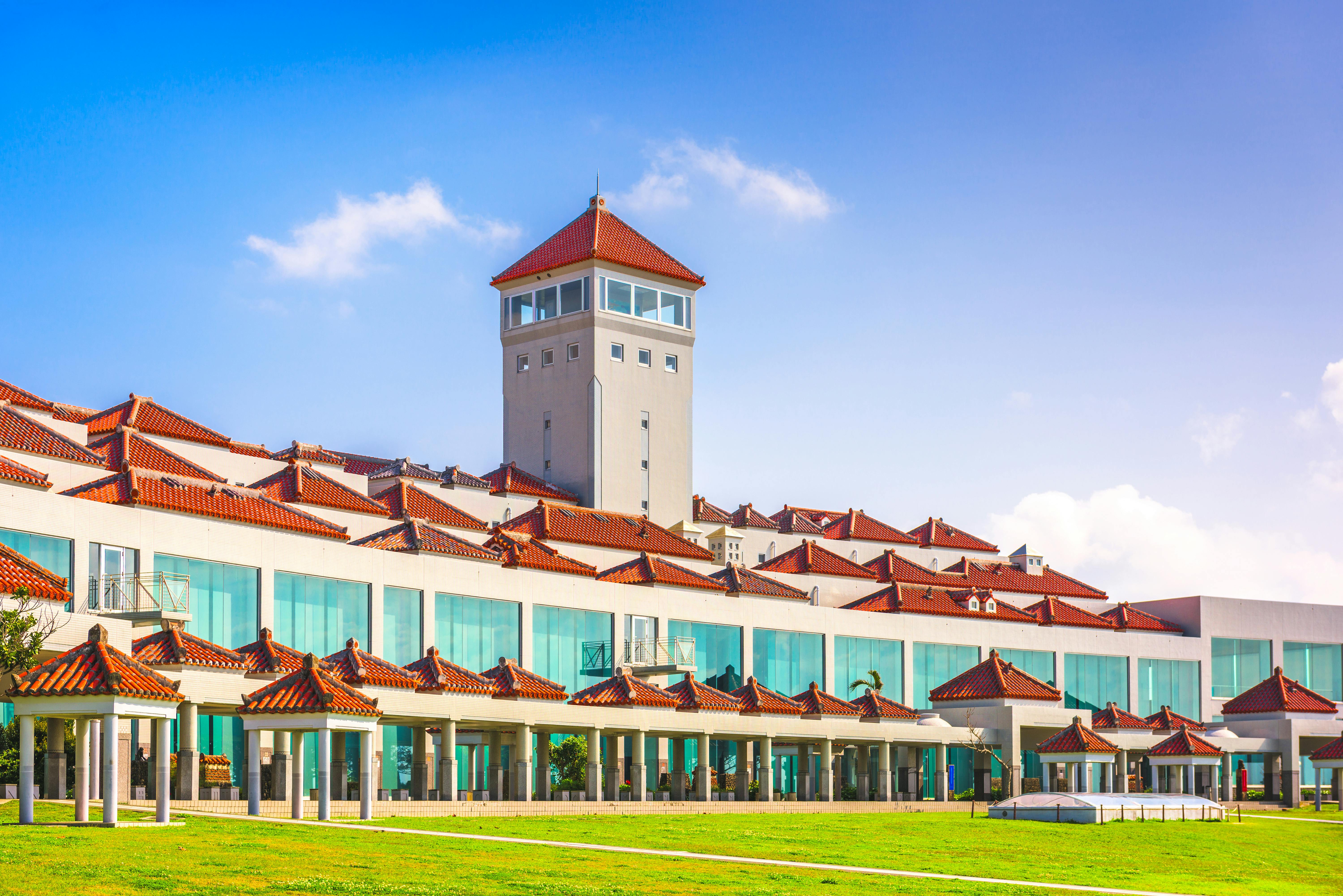 A large modern building with red-tiled roofs, turquoise windows, and a central tower stands under a blue sky, surrounded by green grass and outdoor walkways.