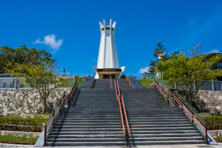 A tall white memorial tower stands at the top of a wide, long staircase with handrails, surrounded by greenery, stone walls, and a bright blue sky above.