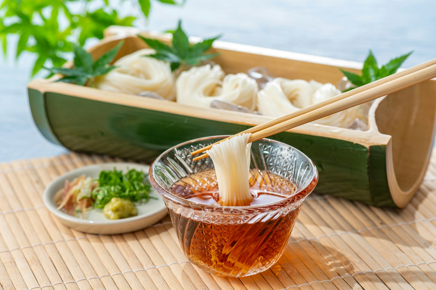 A pair of chopsticks holds thin white noodles dipped in a clear bowl of sauce. Behind, more noodles rest on a bamboo holder with ice cubes. A small plate with ginger, wasabi, and green onions is nearby. The setting includes a bamboo mat and green leaves.