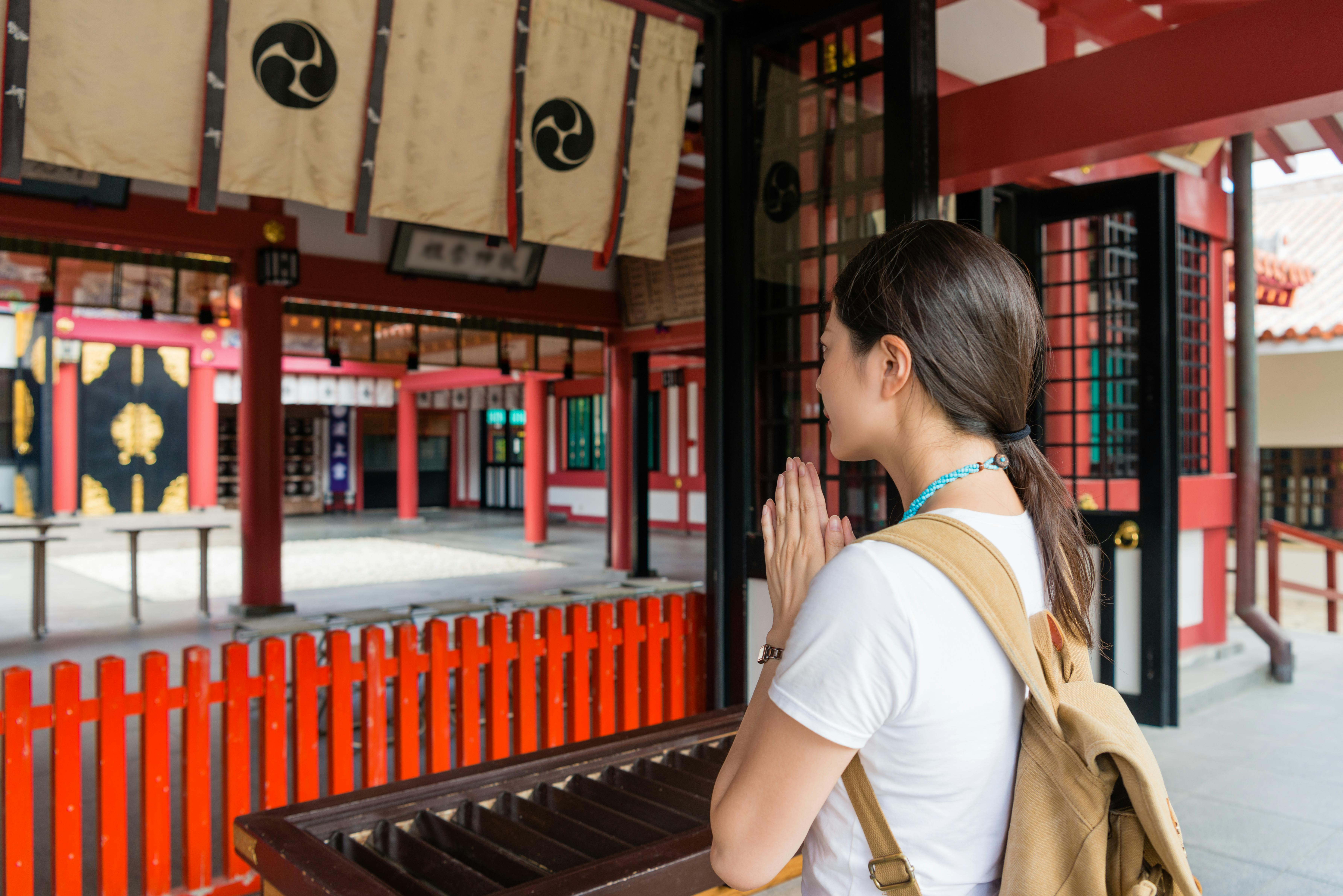 A woman with a backpack stands in front of a Shinto shrine, facing the altar with her hands pressed together in prayer. The shrine features red pillars, wooden structures, and traditional Japanese decorations.