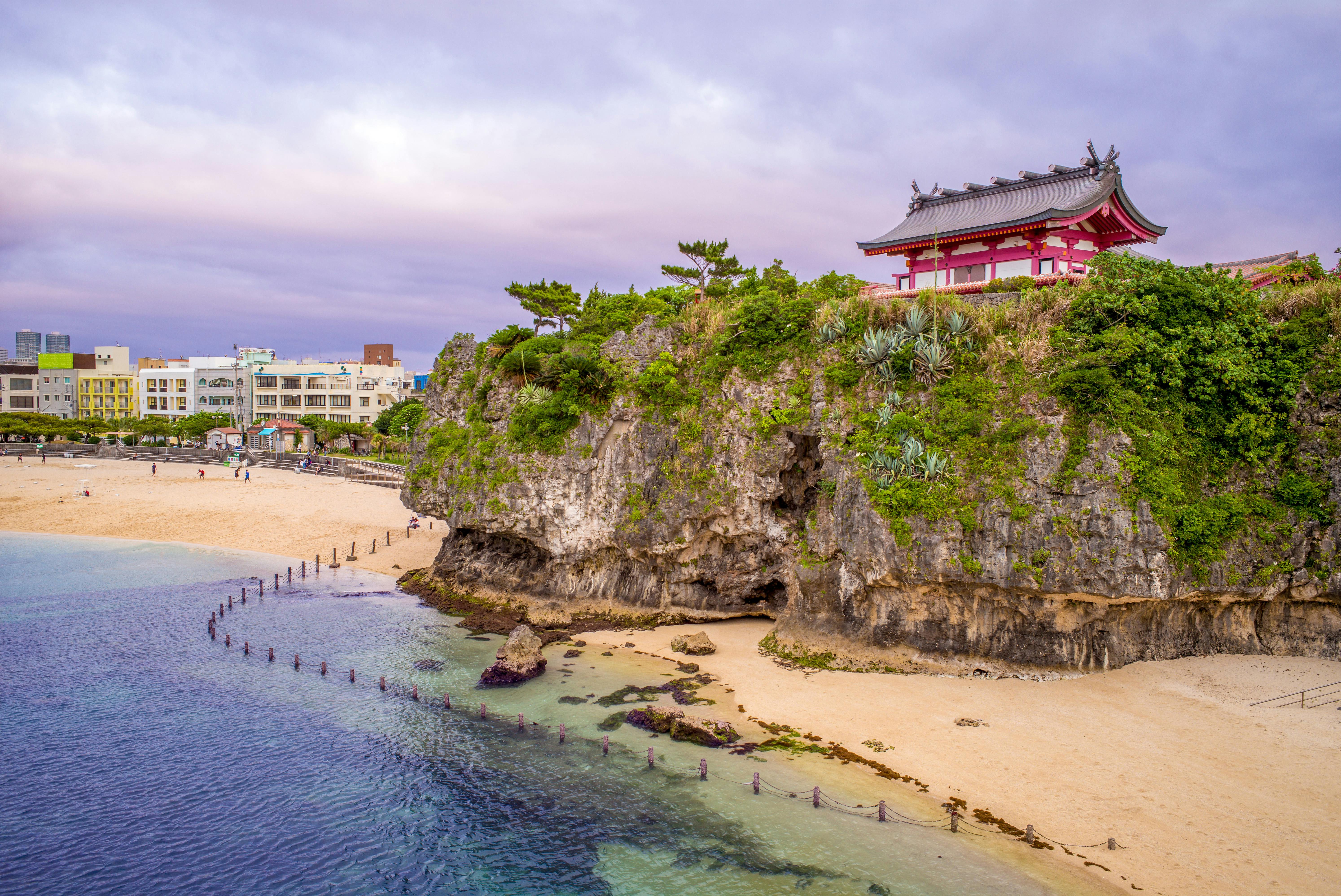 A traditional red and white Japanese shrine sits atop a rocky cliff overlooking a sandy beach and calm blue water, with a cityscape and cloudy sky in the background.