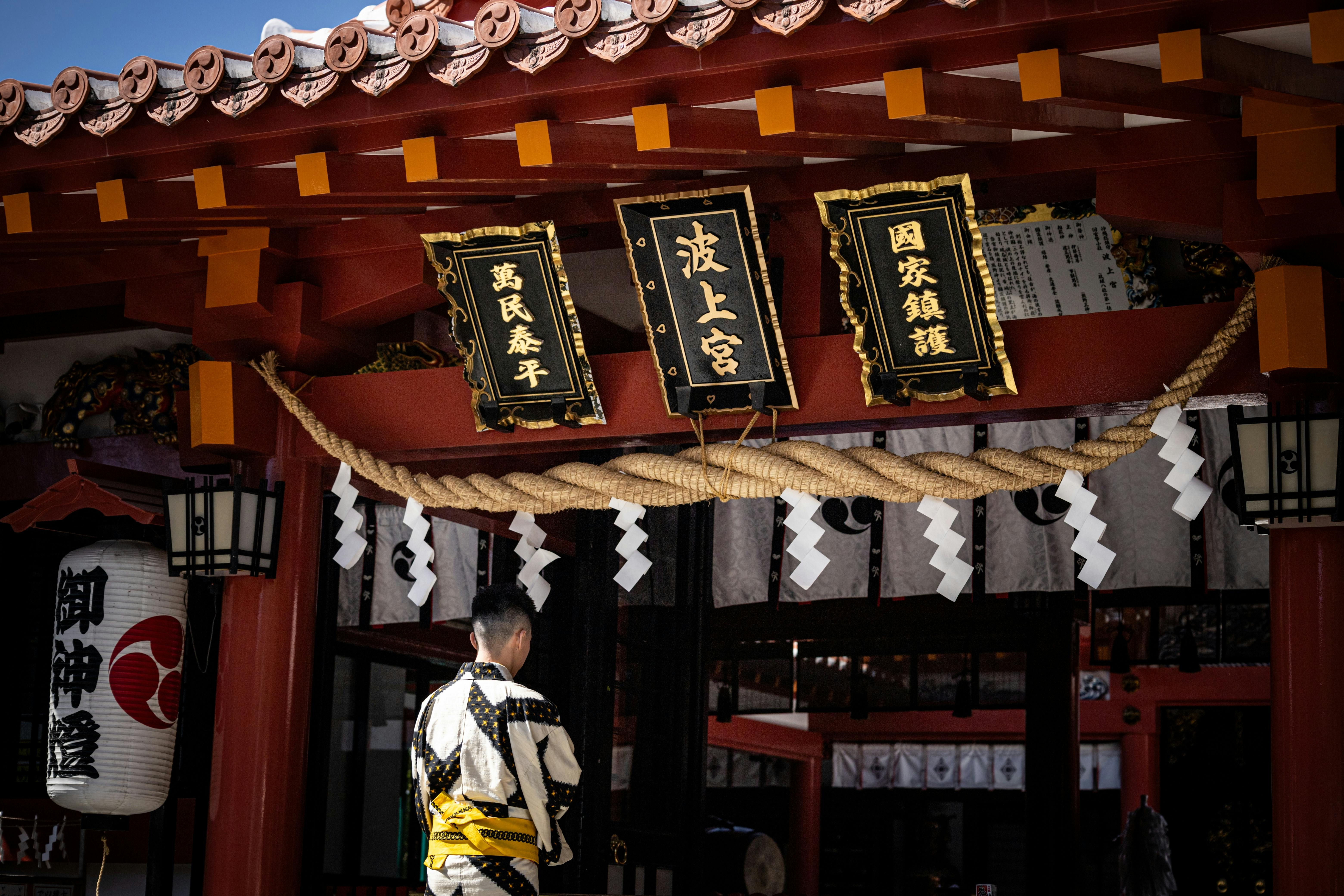 A person in traditional Japanese clothing stands in front of a Shinto shrine with red pillars, hanging lanterns, rope with paper streamers, and three black and gold signs with Japanese writing.
