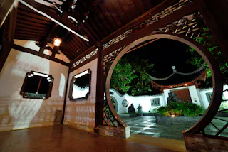 A traditional Chinese garden entrance at night, featuring a round moon gate, ornate woodwork, white walls, and tiled roof. Soft lighting highlights the pathway, plants, and architectural details.