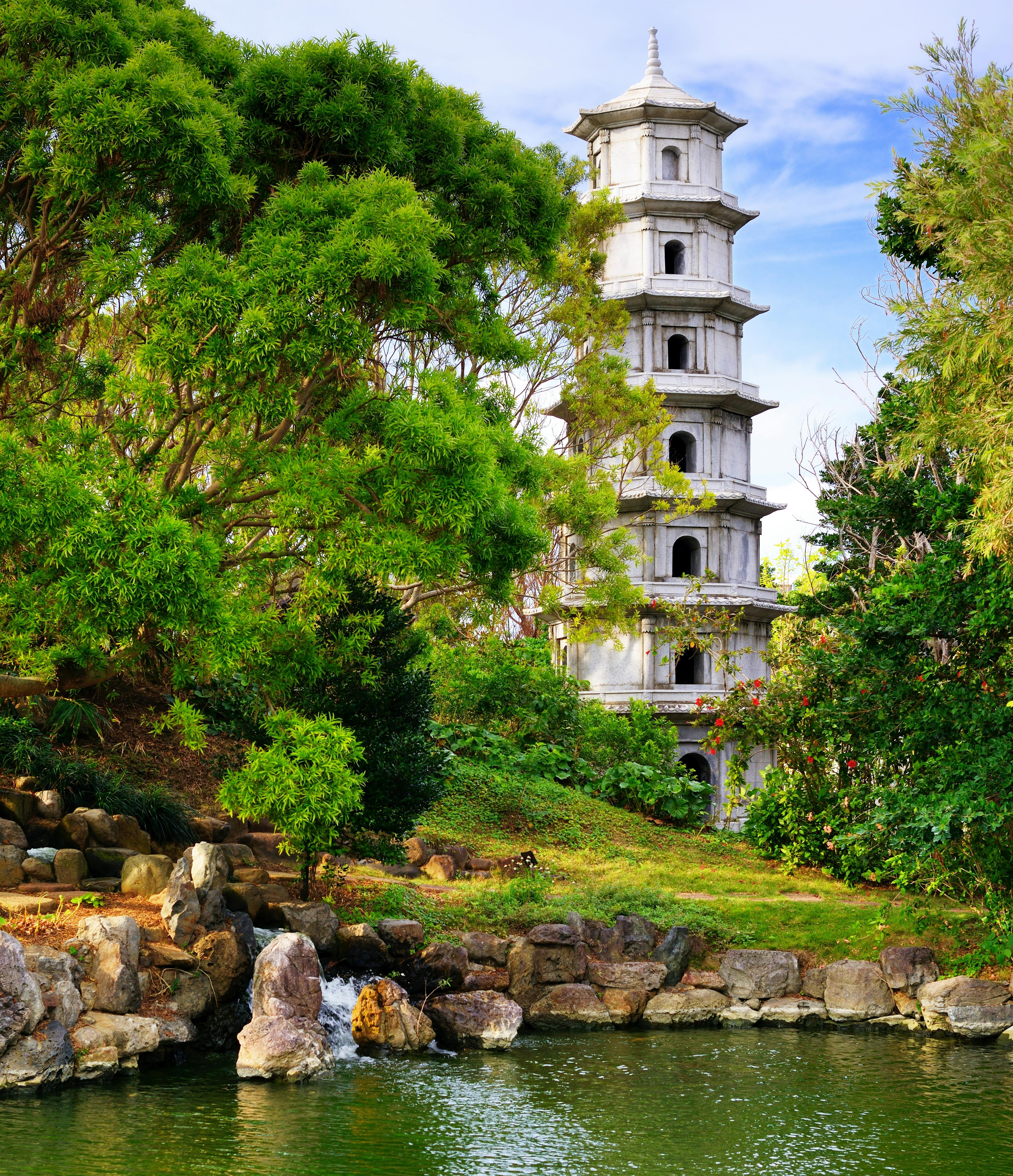A tall, white pagoda rises above green trees and shrubs beside a calm pond with rocks along the shore, under a bright blue sky with scattered clouds.