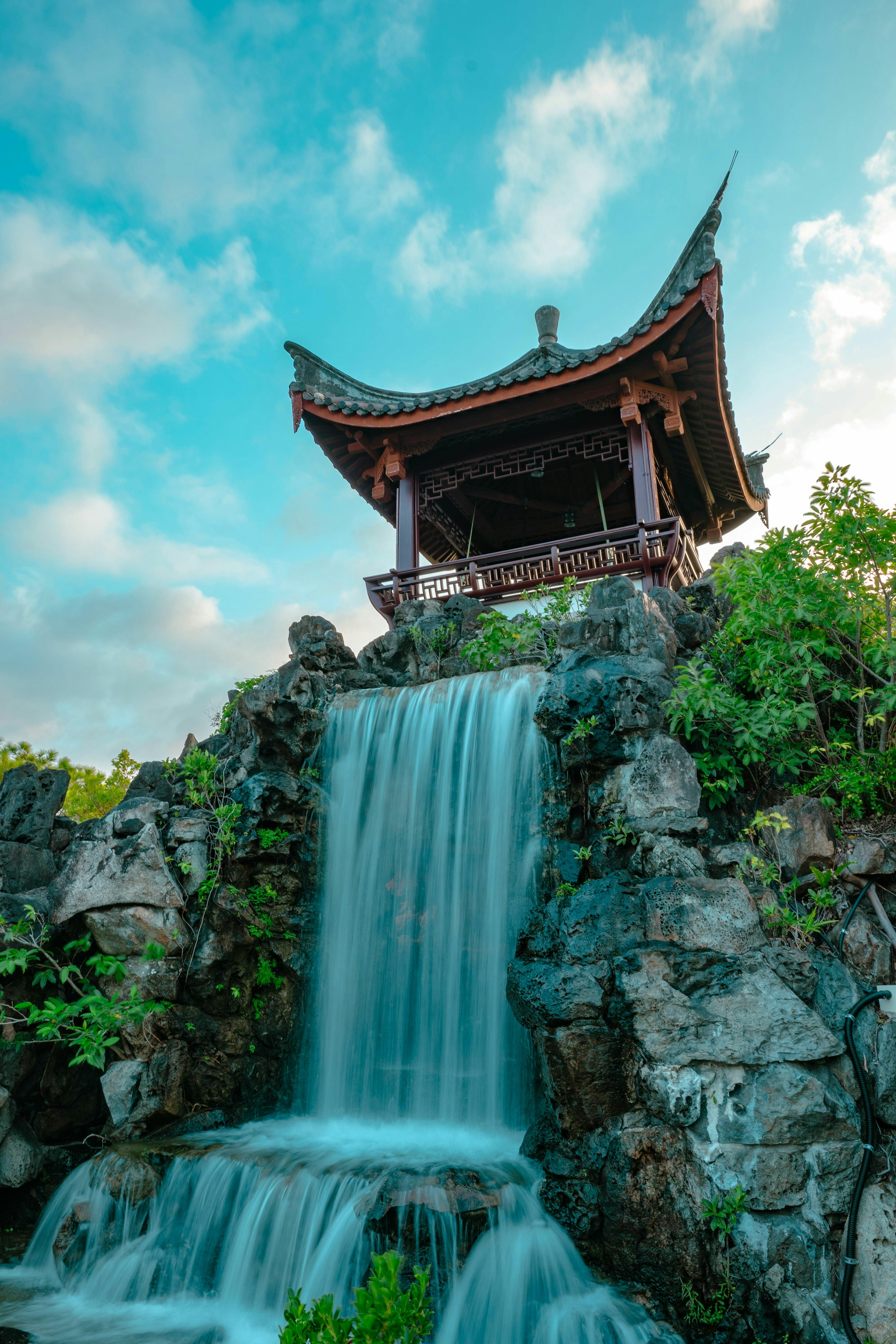 A traditional Asian-style pavilion stands atop a rocky hill with a cascading waterfall flowing beneath it, surrounded by lush green foliage under a blue sky with scattered clouds.