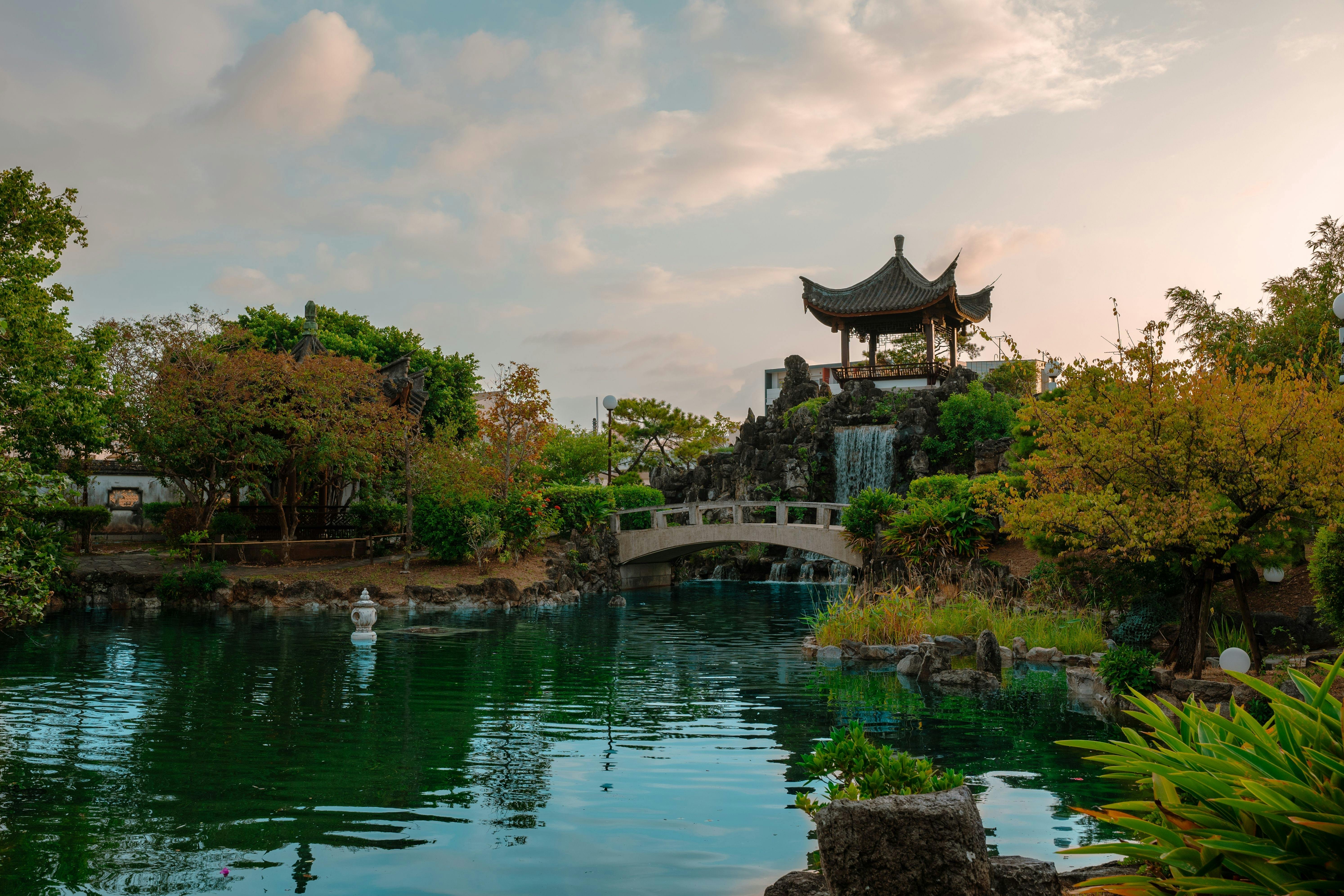 A serene garden with a pond, arched stone bridge, lush greenery, and a traditional pavilion on a small hill with a waterfall, under a partly cloudy sky at sunset.