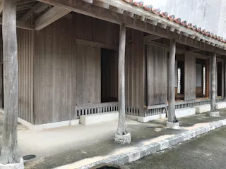 A traditional wooden Japanese building with sliding doors, exposed beams, and a tiled roof. The structure has natural wood pillars and sits on a concrete base with an open porch area.