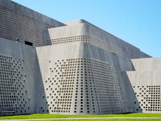 A modern concrete building with sloped walls features a grid of small, square openings, creating a textured geometric pattern; green grass and a blue sky are visible in the background.