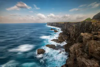 Waves crash against rugged, rocky cliffs under a partly cloudy sky. The ocean appears deep blue, while the rocks and cliffs show rough, natural textures. The scene conveys a sense of dramatic coastal beauty.