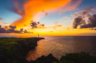 A dramatic sunset sky with vibrant orange and yellow clouds over a rocky coastline, a lighthouse on the cliff, and the calm ocean reflecting the colorful light. Lush green plants are visible in the foreground.