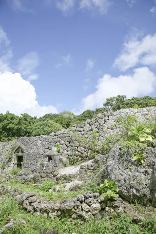 Ancient stone ruins with low walls and a small entryway, surrounded by green vegetation and under a bright blue sky with scattered clouds. Dense trees form the background.