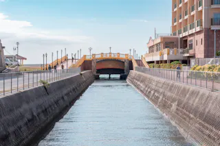 A canal lined with concrete walls leads to a small yellow bridge. Pedestrians walk along paths on both sides, with a multi-story building on the right and clear blue sky overhead.