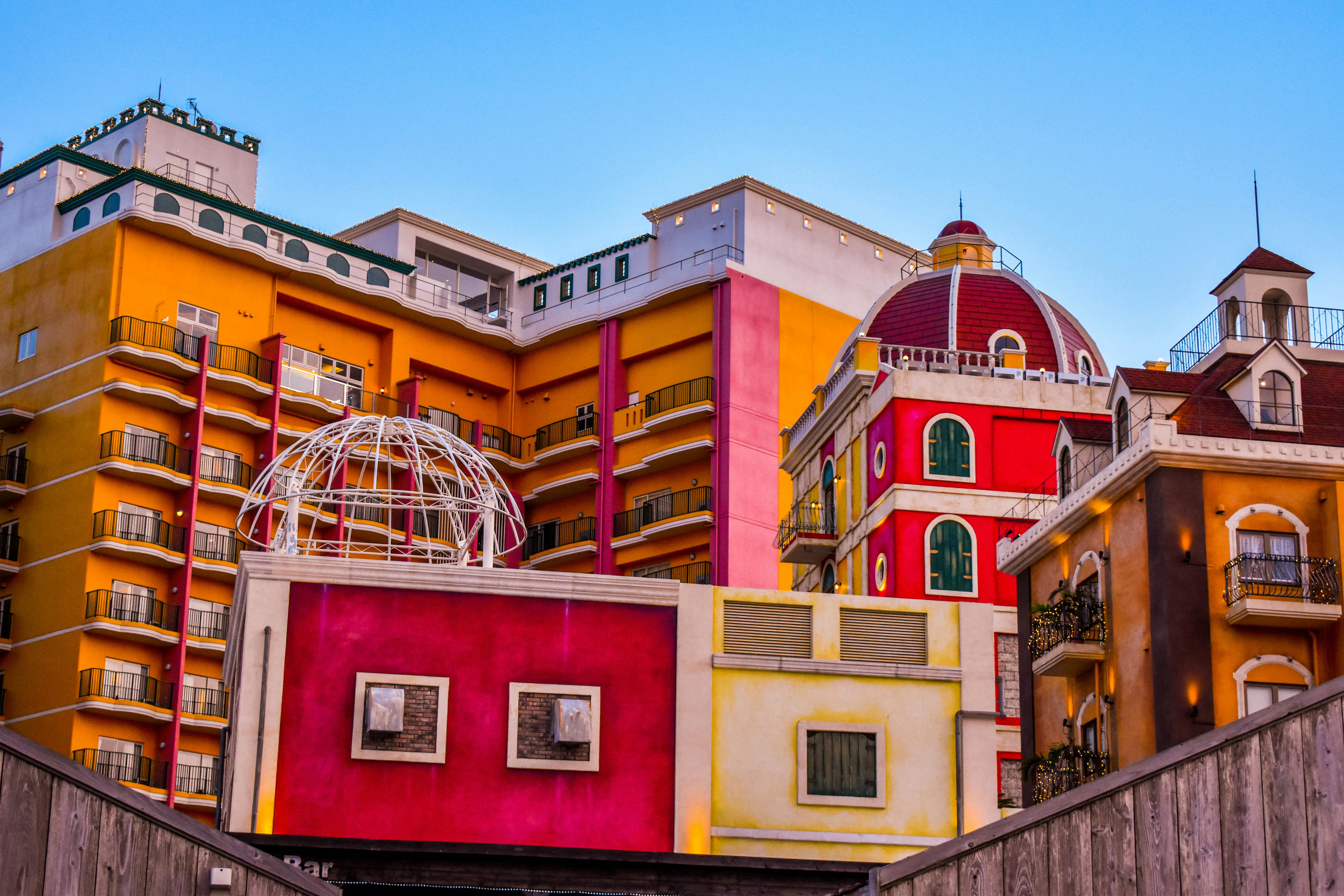 Colorful buildings with vibrant red, yellow, and orange facades, varied balconies, a dome, and a white metal dome structure under a clear blue sky. The architecture is playful and whimsical.