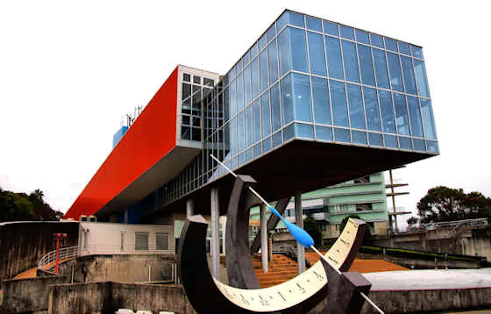 A modern architectural structure with a rectangular glass and orange-accented building elevated on stilts. In the foreground is a large equatorial sundial sculpture with blue accents. The setting appears to be daytime with a cloudy sky.