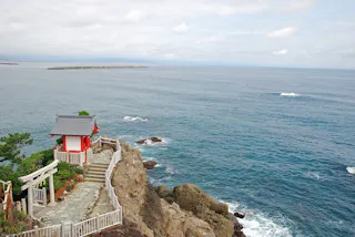 A small red and white shrine sits atop rocky cliffs overlooking a vast blue ocean, with a stone path and torii gate leading to it. The sky is partly cloudy, and distant land and a breakwater are visible on the horizon.
