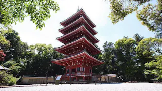 A traditional five-story red pagoda stands surrounded by green trees under a clear blue sky, with sunlight illuminating its intricate wooden architecture.