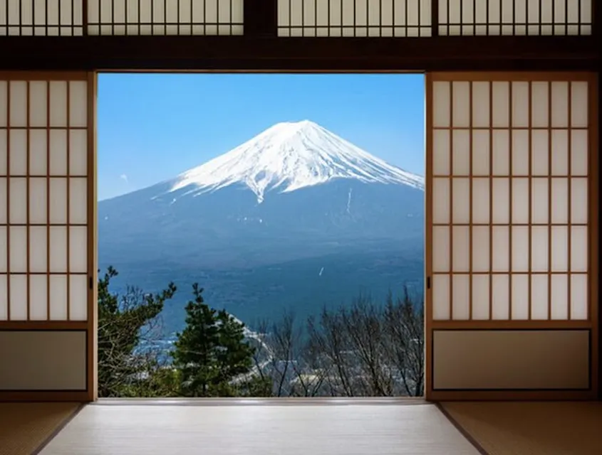 A traditional Japanese room with sliding shoji doors open to reveal a view of Mount Fuji. The mountain is snow-capped under a clear blue sky, framed by the wooden structure and elements of nature like trees.