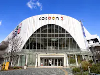 A modern shopping mall with a large arched glass entrance and a curved white facade labeled "Cocoon 2," under a clear blue sky, with trees and plants near the front.
