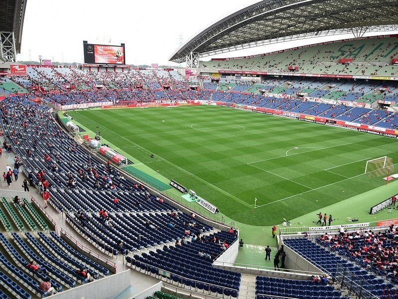 Wide view of a large soccer stadium with green field and blue seats. Some spectators are scattered in the stands, mostly on the far side, and the stadium roof arches overhead. The sky appears overcast.
