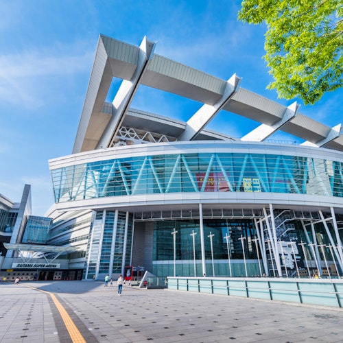 Saitama Super Arena A modern stadium with a distinctive roof design featuring large, white supports. The structure has extensive glass facades and a wide entrance area. The sky is clear, and there are a few people near the entrance and green foliage in the top right corner.