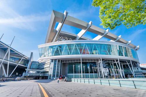 A modern stadium with a distinctive roof design featuring large, white supports. The structure has extensive glass facades and a wide entrance area. The sky is clear, and there are a few people near the entrance and green foliage in the top right corner.