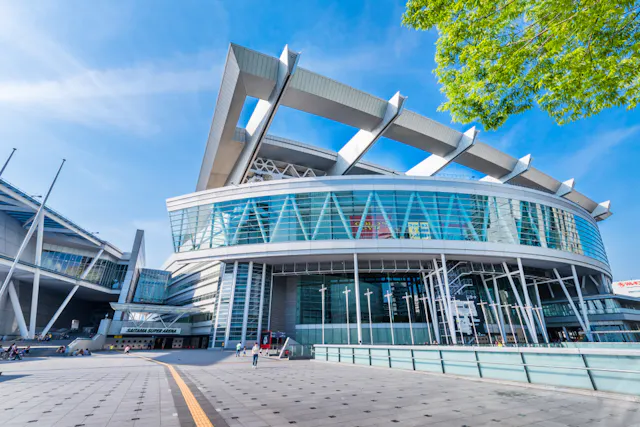 A modern stadium with a distinctive roof design featuring large, white supports. The structure has extensive glass facades and a wide entrance area. The sky is clear, and there are a few people near the entrance and green foliage in the top right corner.