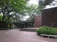 A modern brick building with large windows and stepped entrance, surrounded by trees and greenery. The courtyard features a patterned brick walkway and a concrete bench. Yellow flags are visible near the building.