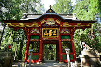 A vibrant red and gold Japanese temple gate stands among tall green trees, flanked by two fox statues on stone pedestals, with intricate carvings and Japanese characters above the entrance.