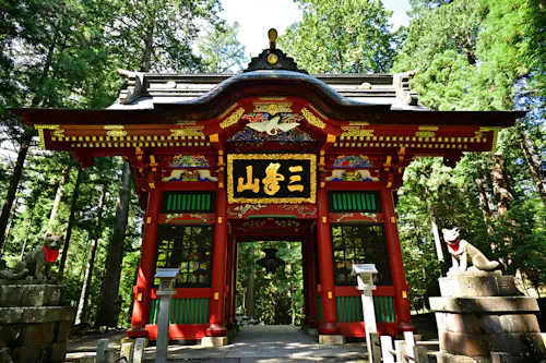 A vibrant red and gold Japanese temple gate stands among tall green trees, flanked by two fox statues on stone pedestals, with intricate carvings and Japanese characters above the entrance.