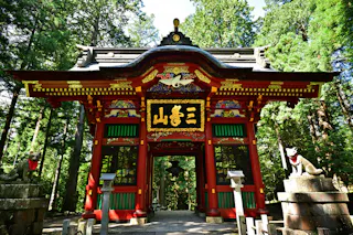 A vibrant red and gold Japanese temple gate stands among tall green trees, flanked by two fox statues on stone pedestals, with intricate carvings and Japanese characters above the entrance.