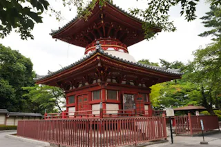 A traditional Japanese pagoda with red wooden beams and ornate roof details stands surrounded by green trees and a red fence on a sunny day.