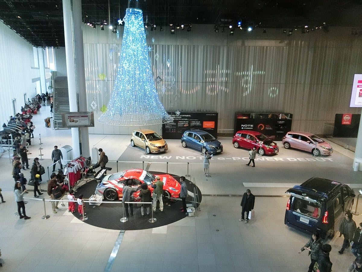 A spacious car showroom with several cars on display, people walking around, and a large illuminated tree-shaped decoration hanging from the ceiling. The floor has the words "NEW NOTE e-POWER DEBUT" written on it.