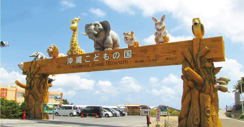 Entrance gate of Okinawa Zoo & Museum decorated with large animal statues, including a giraffe, elephant, tiger, rabbit, and monkey, against a blue sky with some buildings in the background.