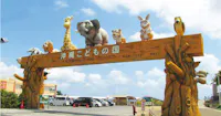 Entrance gate of Okinawa Zoo & Museum decorated with large animal statues, including a giraffe, elephant, tiger, rabbit, and monkey, against a blue sky with some buildings in the background.