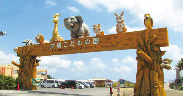 Entrance gate of Okinawa Zoo & Museum decorated with large animal statues, including a giraffe, elephant, tiger, rabbit, and monkey, against a blue sky with some buildings in the background.