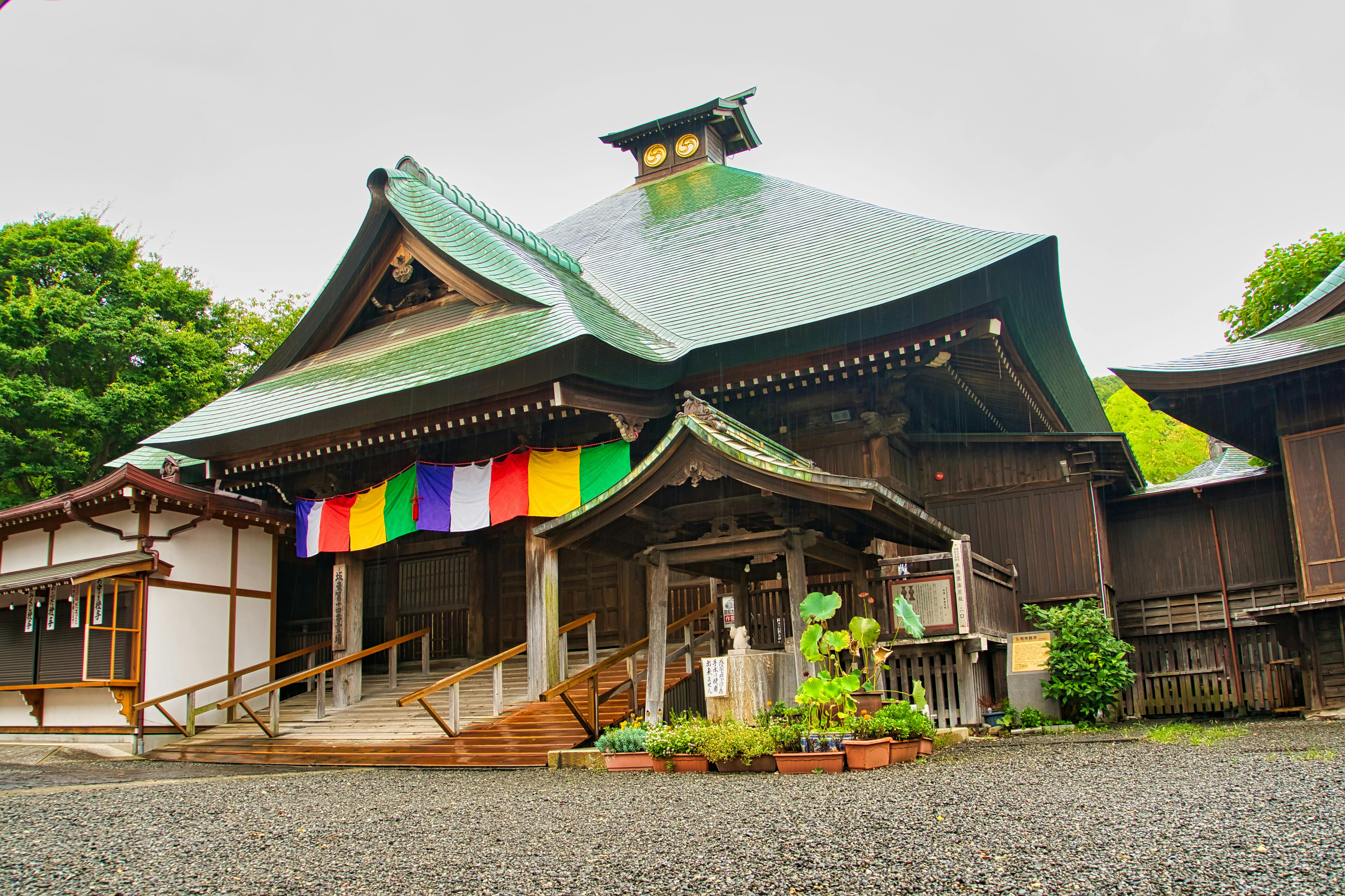 A traditional Japanese shrine with a green tiled roof, colorful hanging banners, wooden architecture, and potted plants at the entrance, surrounded by greenery and a gravel courtyard.