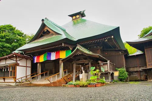 A traditional Japanese shrine with a green tiled roof, colorful hanging banners, wooden architecture, and potted plants at the entrance, surrounded by greenery and a gravel courtyard.