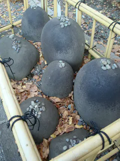 Large smooth stones with small pebbles stacked on top are arranged in a rectangular bamboo enclosure, surrounded by dry fallen leaves and tied at the corners with black cord.