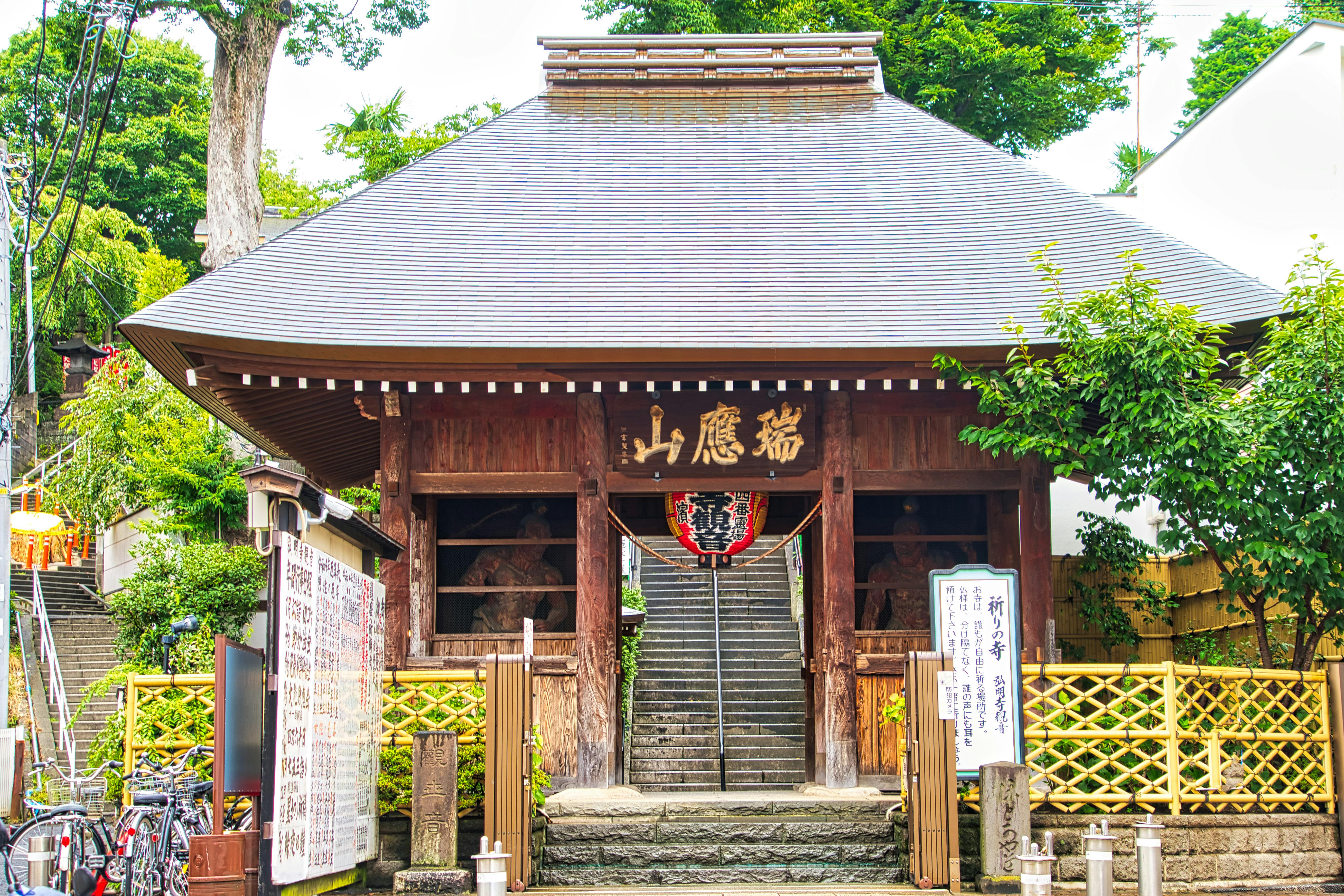 A traditional Japanese temple with a sloped roof, wooden structure, and large gold kanji characters above the entrance. Stone steps lead inside, and greenery surrounds the building. Signs and bicycles are visible nearby.