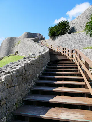 Wooden stairs ascend alongside ancient stone walls of a historic fortress under a bright blue sky, with palm-like trees visible at the top and sunlight casting shadows on the steps.