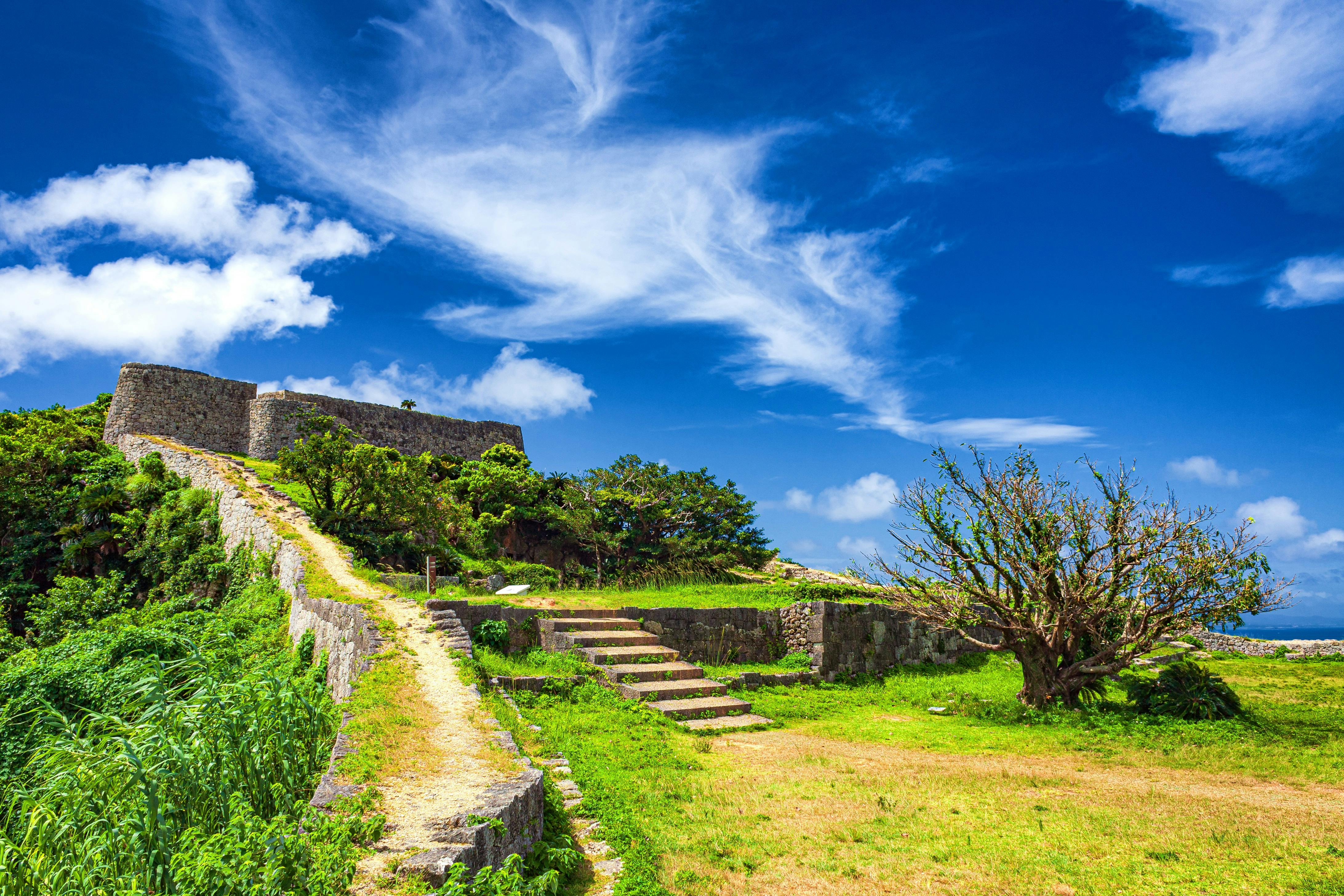 A stone path leads up to the ruins of an ancient stone wall surrounded by lush greenery, under a bright blue sky with wispy white clouds. A large tree stands on the right, basking in sunlight.