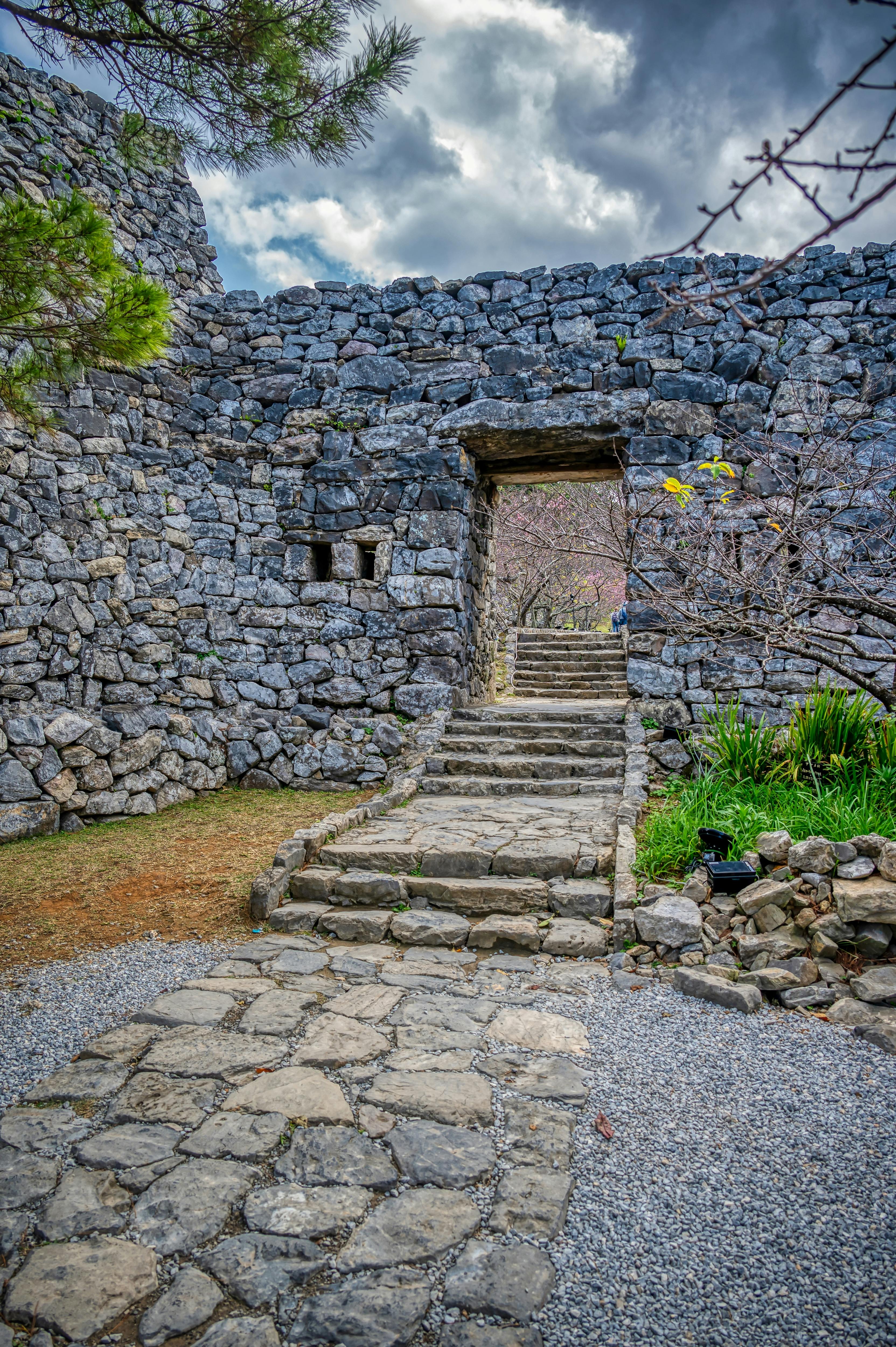 A stone pathway with steps leads through an arched doorway in an ancient stone wall, surrounded by greenery and trees under a cloudy sky.