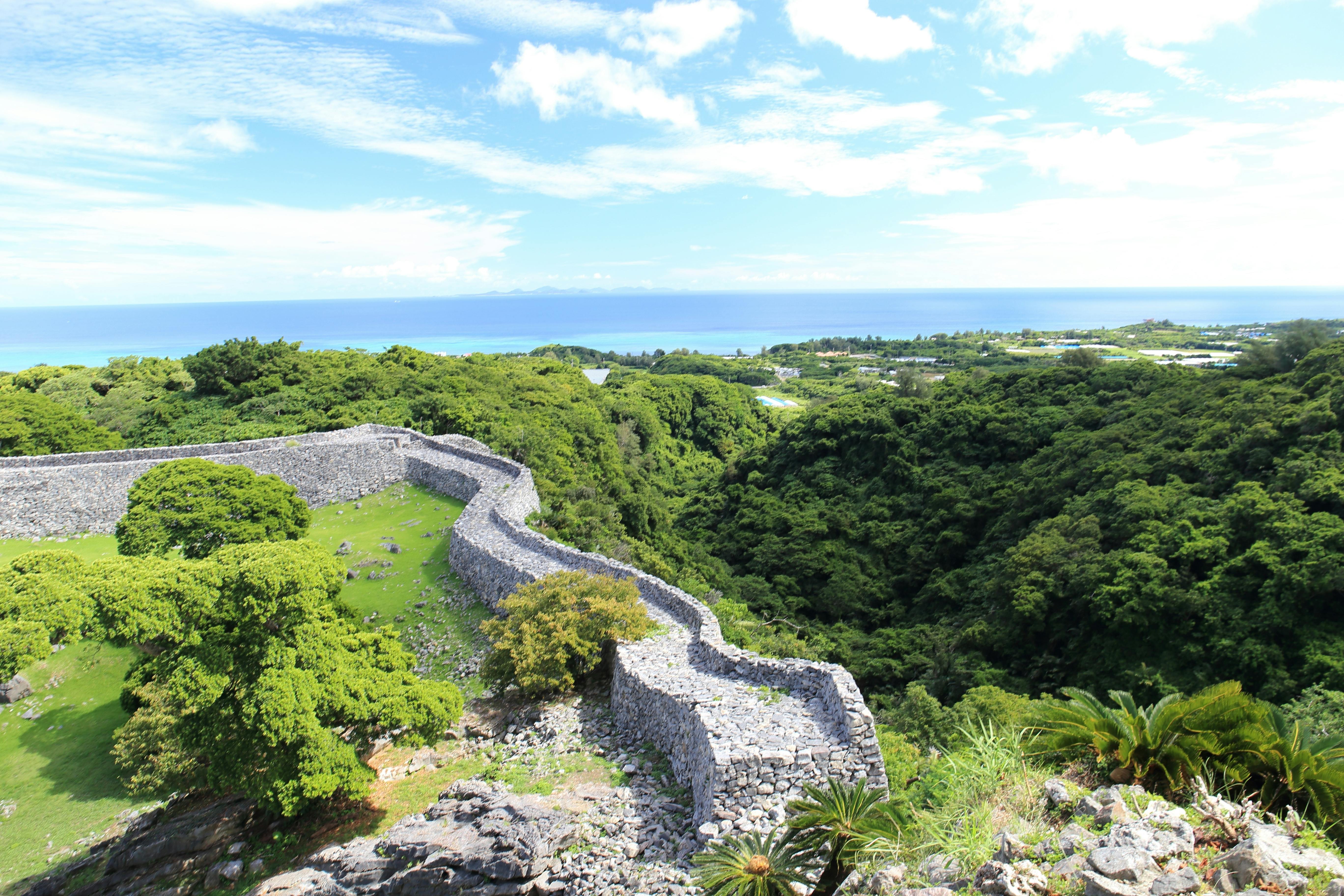 A stone wall winds through lush green trees on a hillside, overlooking a distant coastline and blue ocean under a partly cloudy sky.