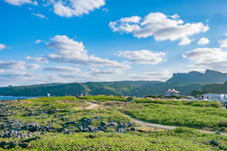 A scenic landscape featuring a green, rocky field with a winding path, distant hills, a small pavilion, and a bright blue sky dotted with white clouds.