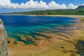 A scenic coastline features clear blue-green water, shallow reefs, and a sandy shore, bordered by lush green hills under a partly cloudy sky.