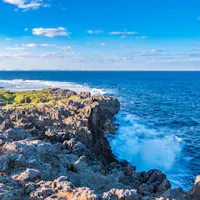 Hedo Misaki Rocky coastline with waves crashing against the jagged rocks, lush greenery on the left, and a vast blue ocean stretching to the horizon under a bright sky with scattered clouds.