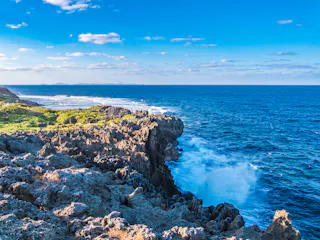 Rocky coastline with waves crashing against the jagged rocks, lush greenery on the left, and a vast blue ocean stretching to the horizon under a bright sky with scattered clouds.