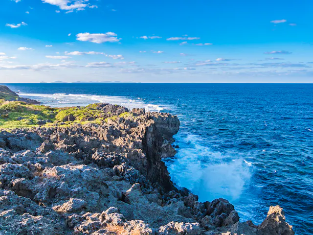 Rocky coastline with waves crashing against the jagged rocks, lush greenery on the left, and a vast blue ocean stretching to the horizon under a bright sky with scattered clouds.