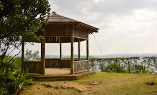 A wooden gazebo with a hexagonal roof stands on a grassy hilltop, surrounded by greenery and overlooking a distant cityscape under a cloudy sky.