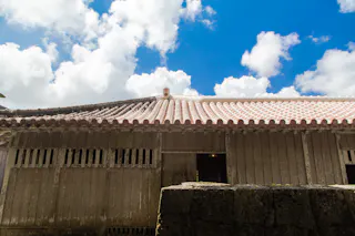 A traditional wooden Japanese house with a tiled roof sits under a bright blue sky filled with scattered white clouds. A low stone wall runs along the foreground.